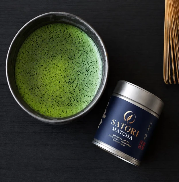 A bowl of whisked matcha beside a SATORI ceremonial matcha tin, seen from above
